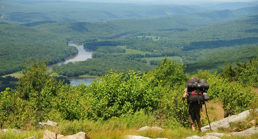 A person wearing a backpack hikes through a green area above a vast green landscape with a river winding through it